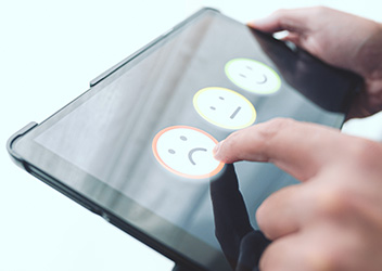A person using a tablet with a feedback interface displaying three emojis: a happy face, a neutral face, and a sad face. The sad face is being selected.
